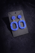 A pair of chunky, glittery blue drop earrings with circular studs and elongated oval dangles, displayed on a black card against a dark textured background.
