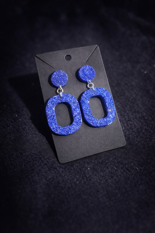 A pair of chunky, glittery blue drop earrings with circular studs and elongated oval dangles, displayed on a black card against a dark textured background.